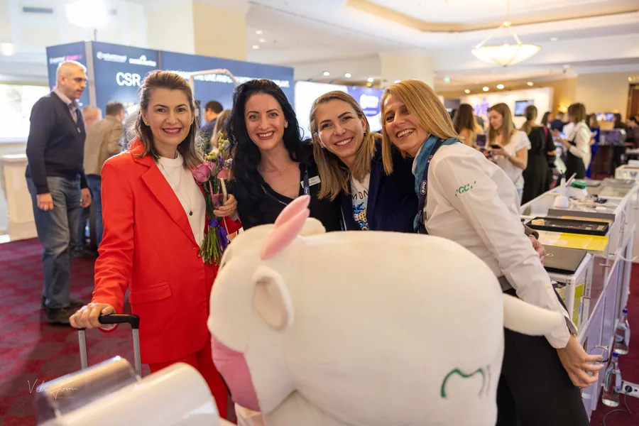 Patru doctorițe zâmbesc lângă o mascotă de pluș în formă de dinte, la un stand din expoziție.